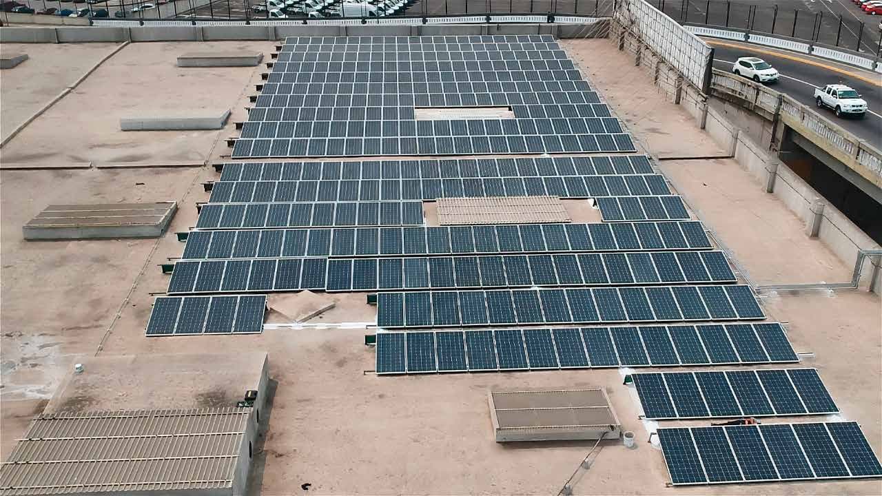 Azotea de un edificio comercial con varias filas de paneles solares sobre estructura, junto a una avenida.