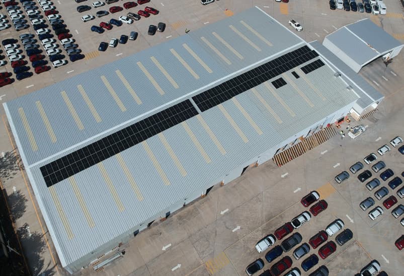 Vista aérea de una nave industrial con dos franjas de paneles solares sobre la cubierta, rodeada de un estacionamiento.